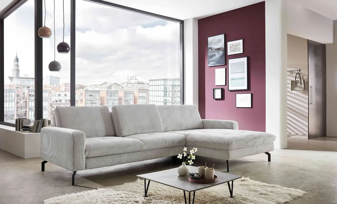 Helles Ecksofa vor bodentiefen Fenstern, niedriger Couchtisch mit Tablett und Vase, bordeauxrote Wand mit mehreren gerahmten Bildern im Hintergrund.